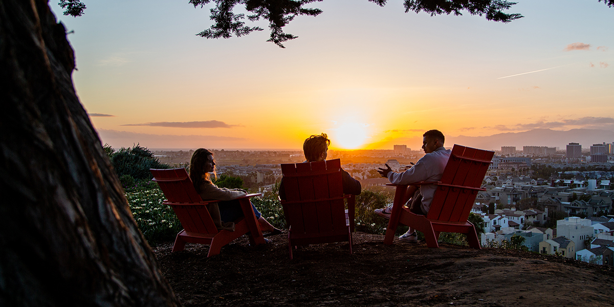 Three individuals sitting in chairs on the bluff at sunset with their backs to the camera.